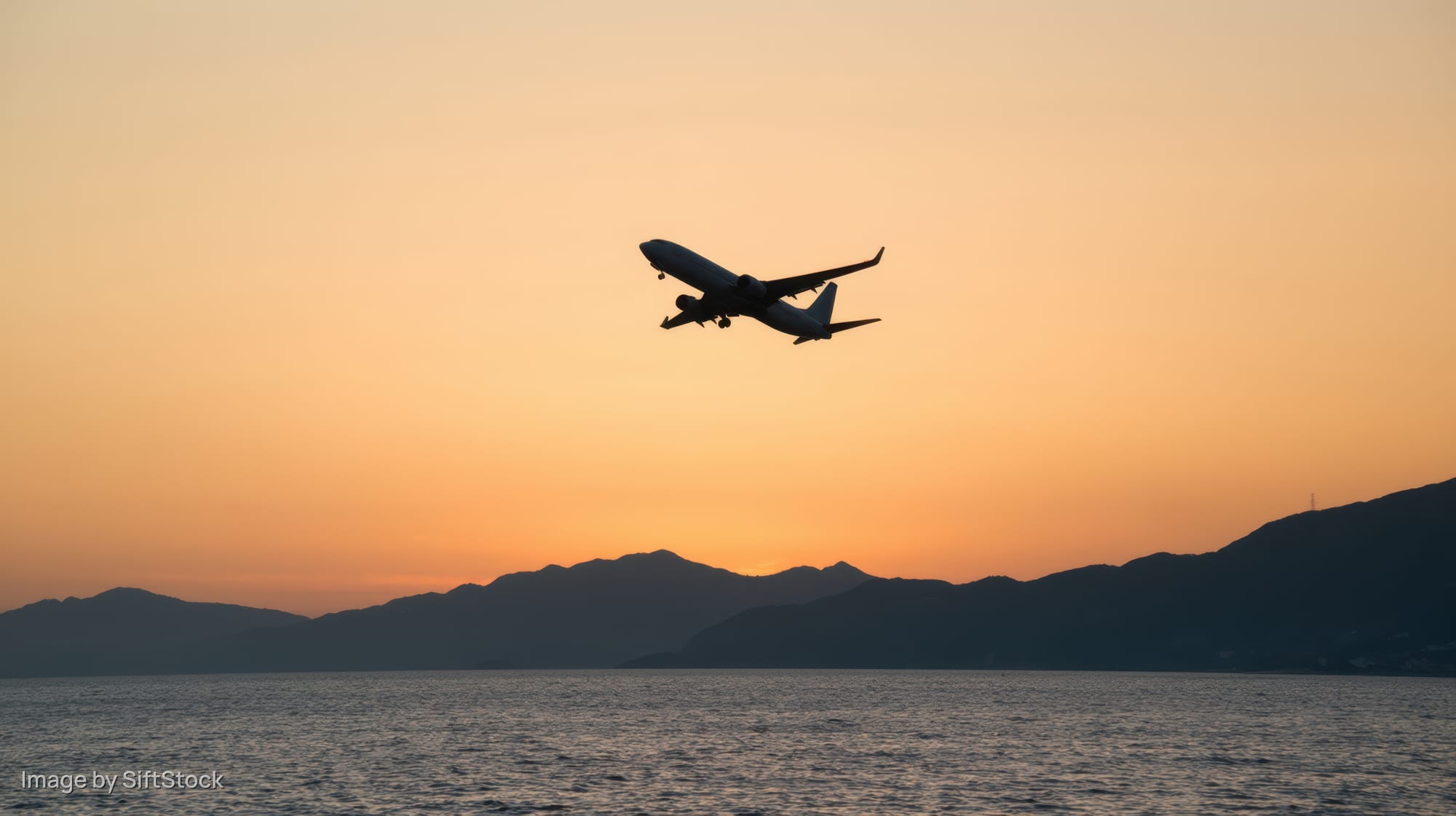 Airplane silhouetted against sunset sky flying low over ocean with mountains in background