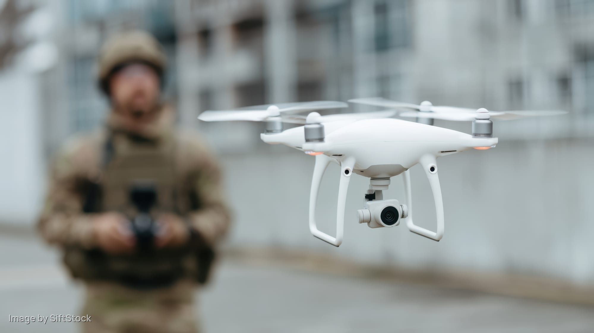 White quadcopter drone hovering in front of a soldier in urban environment