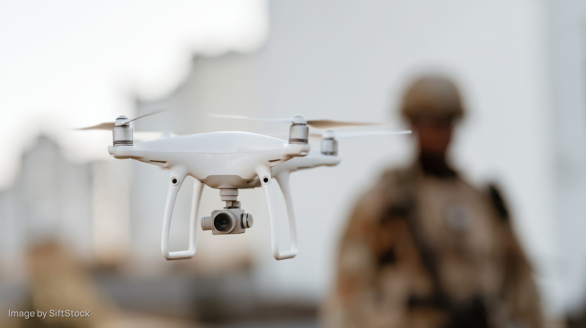 White quadcopter drone hovering in front of a soldier in desert environment