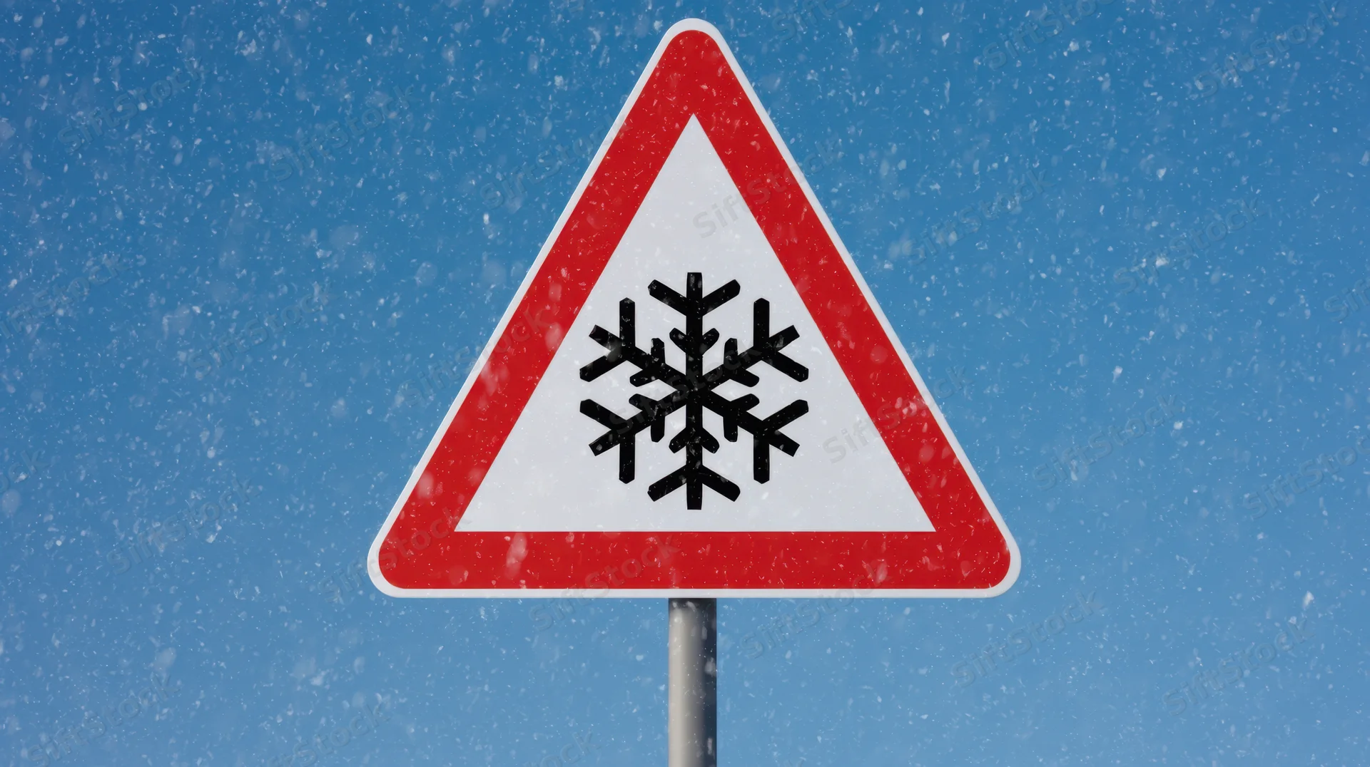 Triangular warning sign with snowflake symbol against a blue sky during snowfall