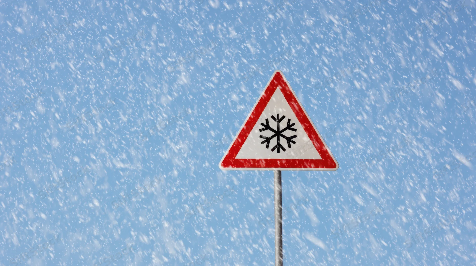 Triangular warning sign with snowflake symbol against blue sky during snowfall