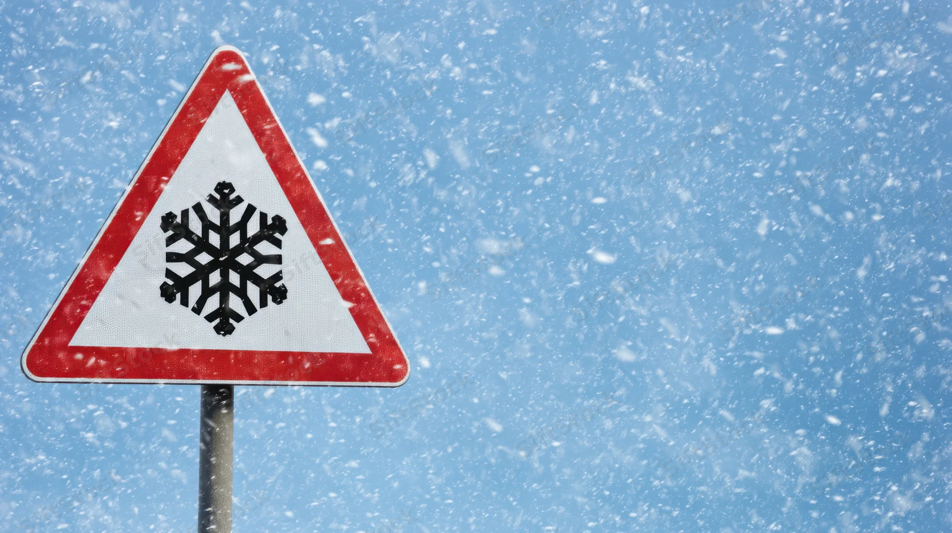 Triangular warning sign with snowflake symbol against a blue sky during snowfall