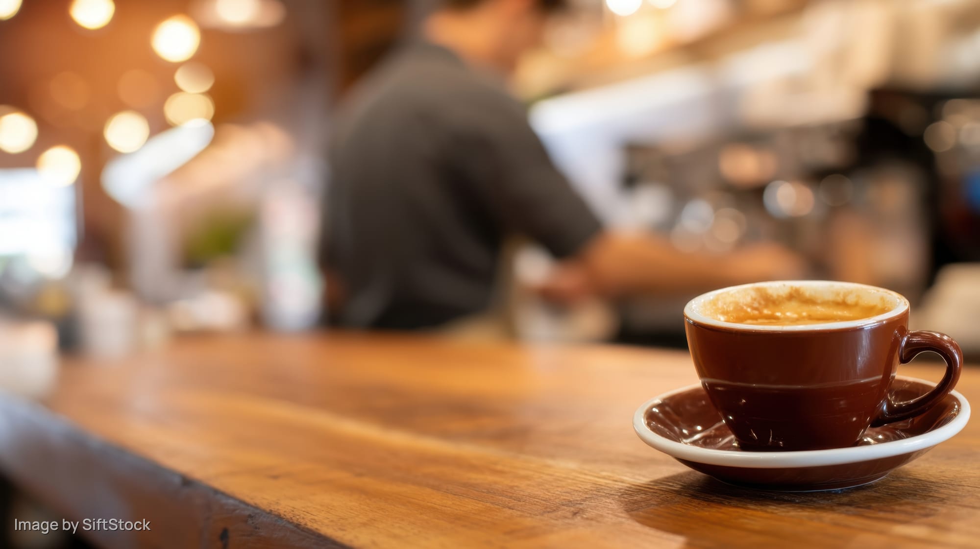 Brown ceramic coffee cup on wooden table in warm cafe interior with barista in background