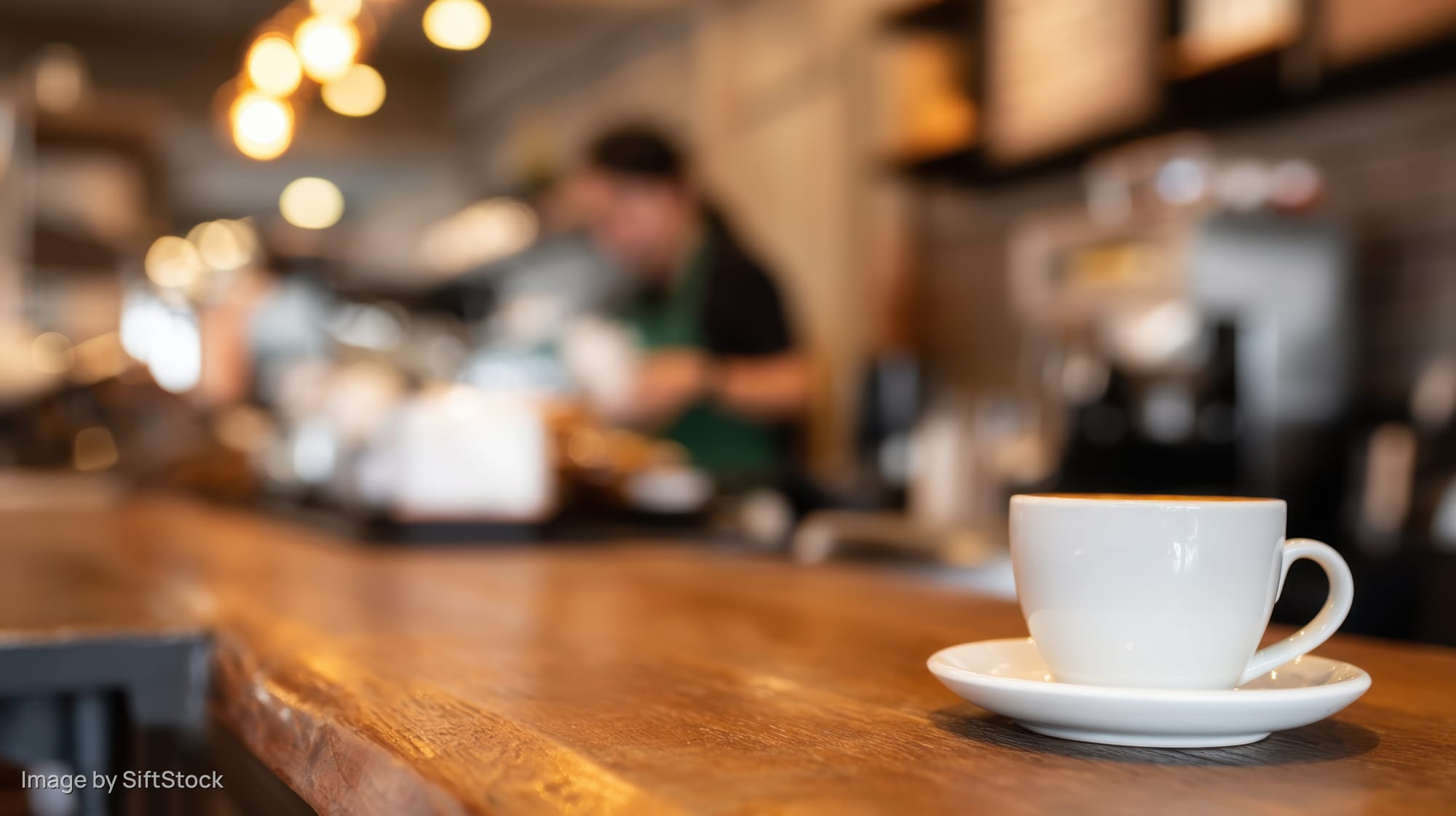 White coffee cup on wooden counter in warm cafe interior with barista in background