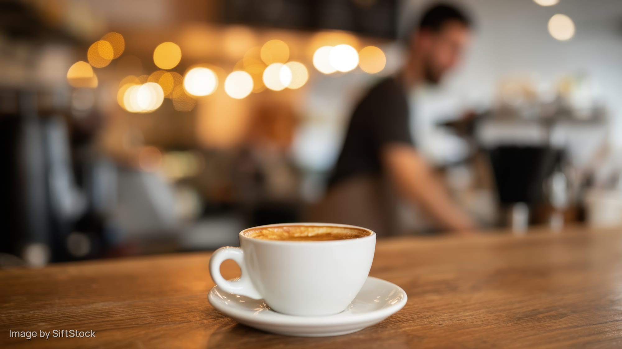 White coffee cup with foam on wooden table in cafe interior with blurred barista and warm lights in background
