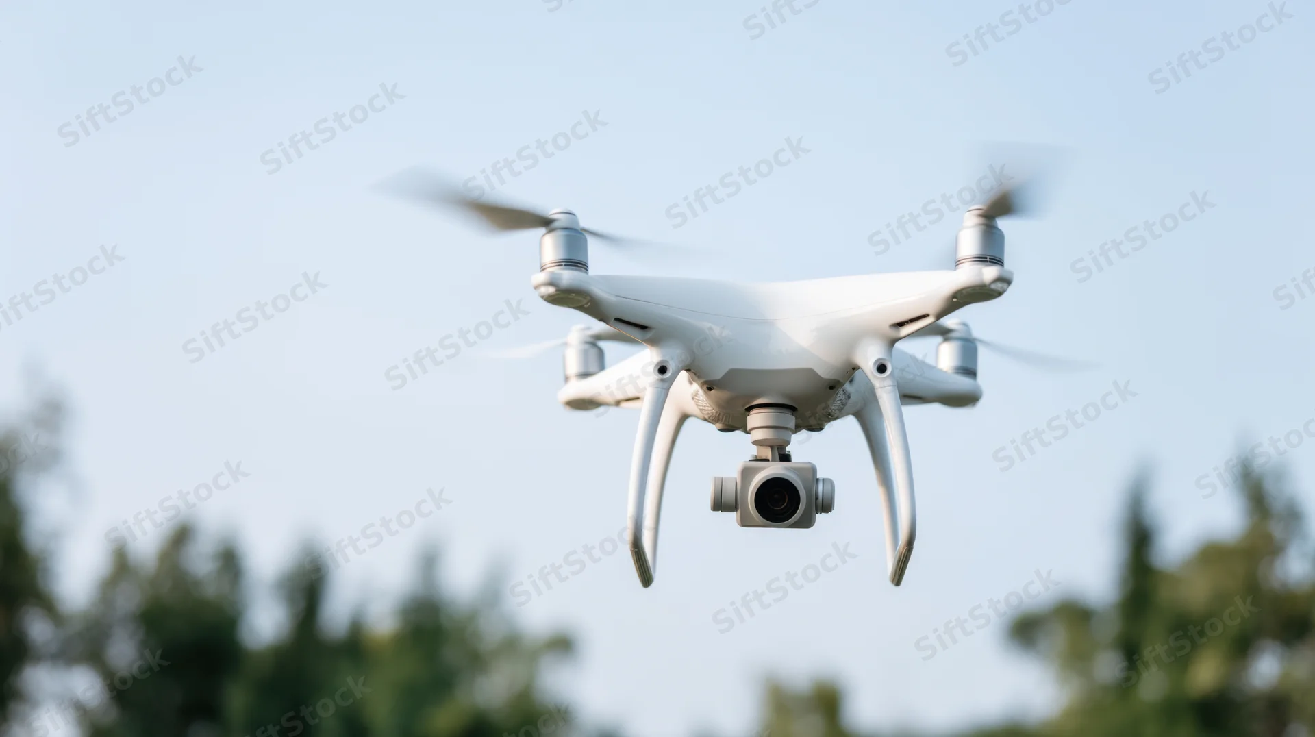 White quadcopter drone hovering in sky with camera and spinning propellers above trees