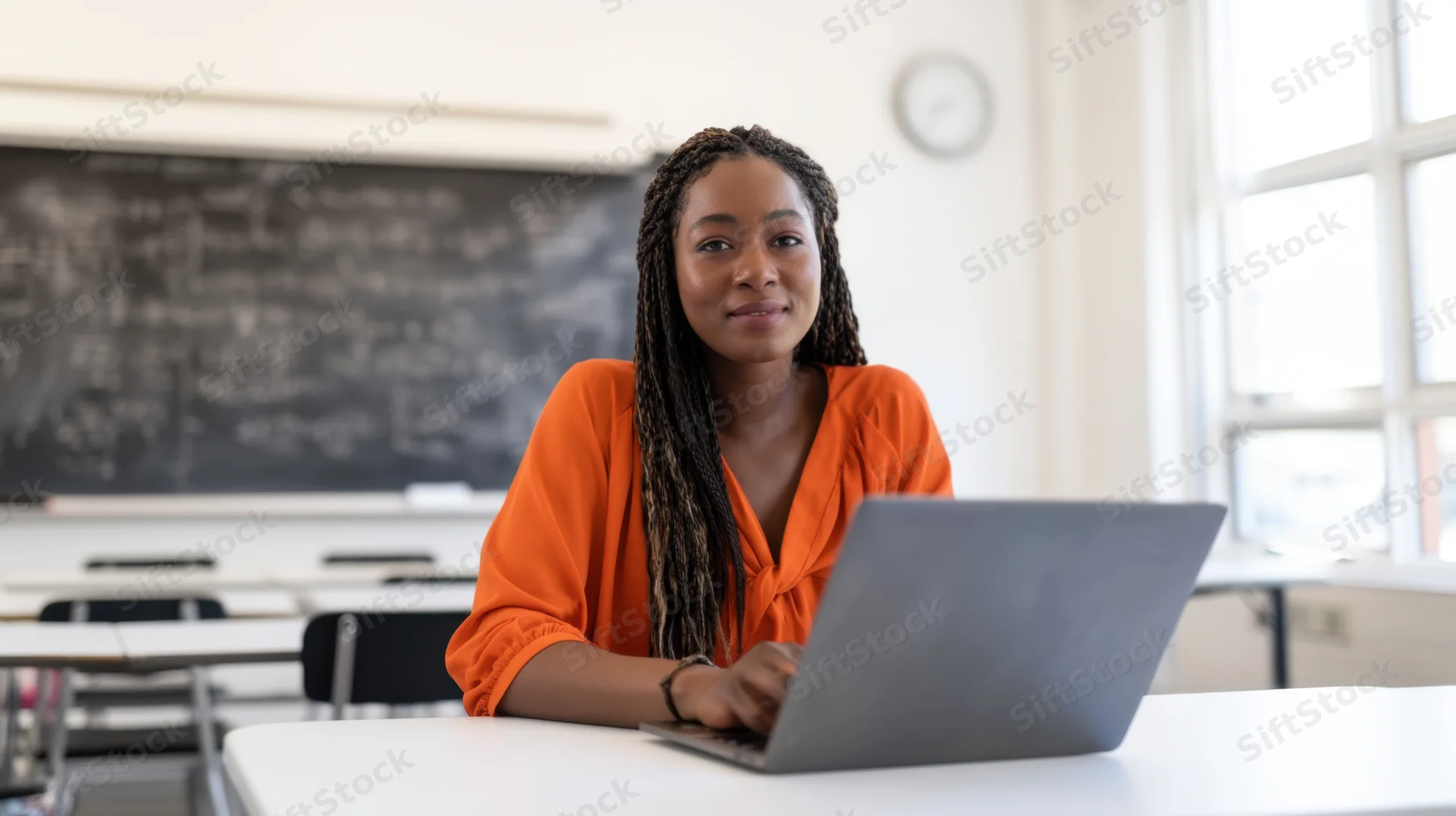 Woman with braided hair smiling while using laptop at desk in classroom