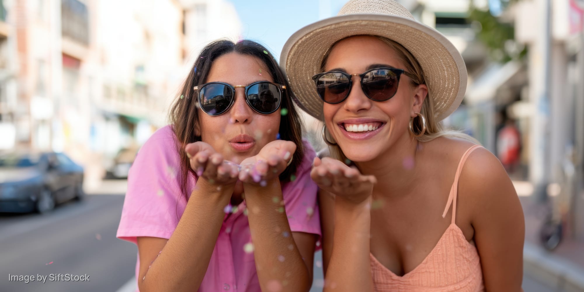 Two women wearing sunglasses blowing confetti on a city sidewalk