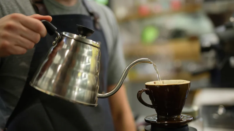 Barista pouring hot water from a gooseneck kettle into a ceramic coffee filter in a cafe