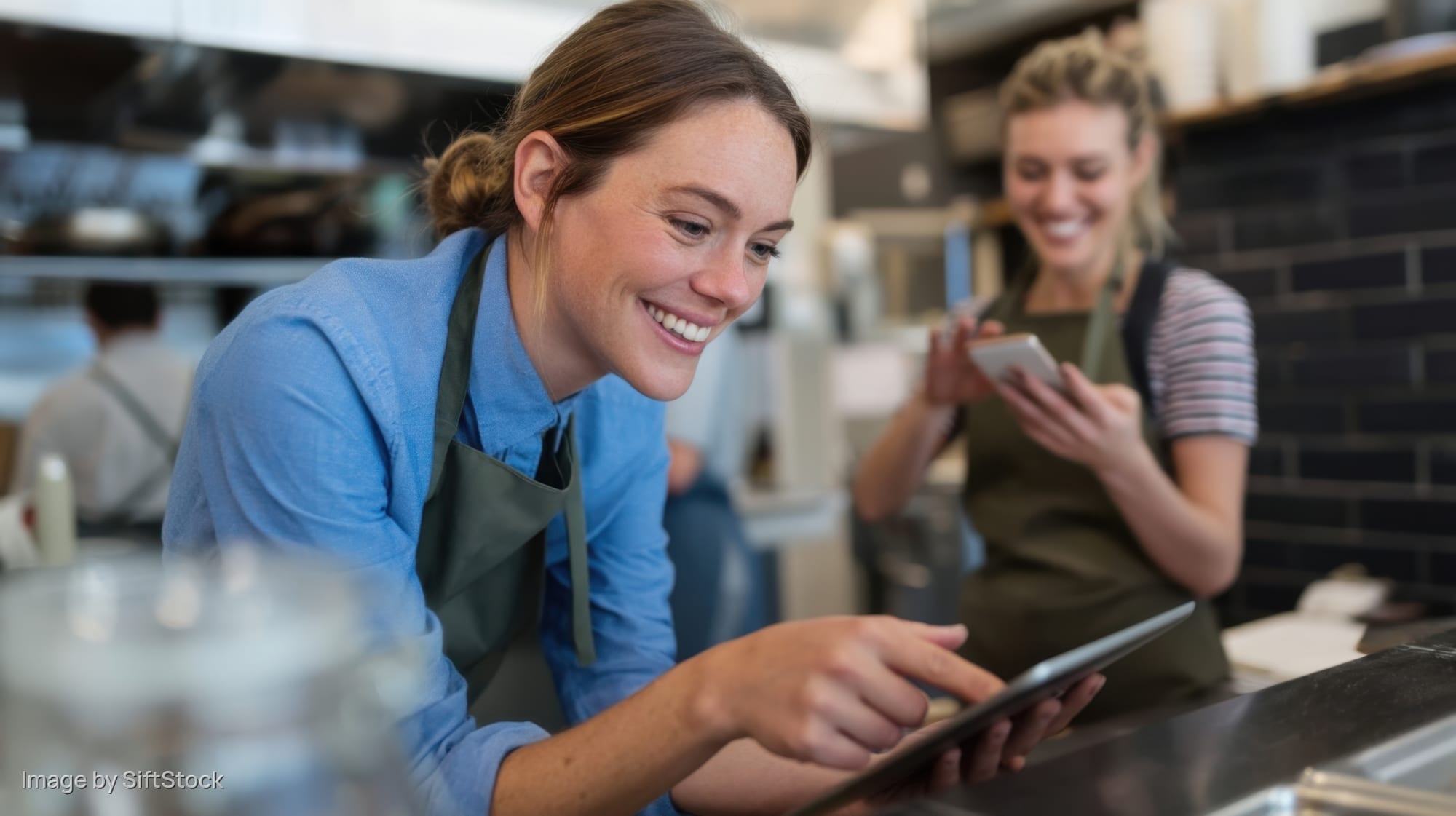 Woman barista smiling while using tablet at cafe counter with colleague in background