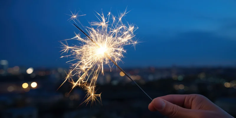 Hand holding lit sparkler against city skyline at twilight
