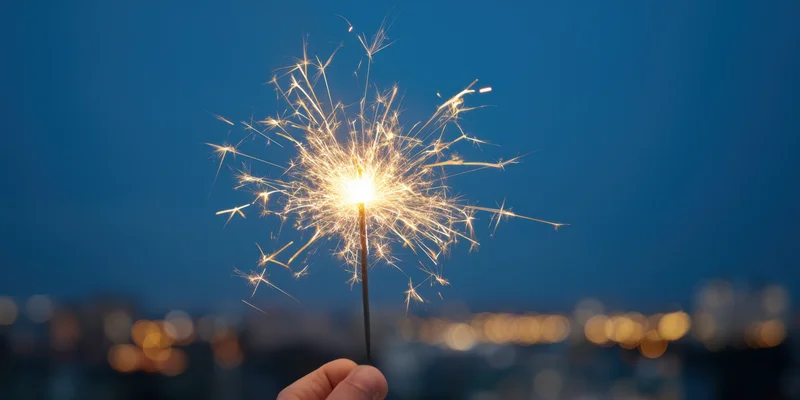Hand holding lit sparkler against city lights at night