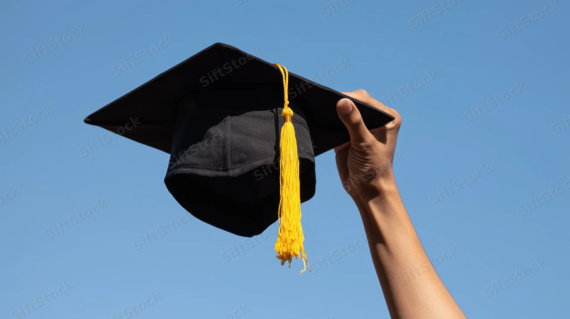 Hand holding black graduation cap with yellow tassel against blue sky