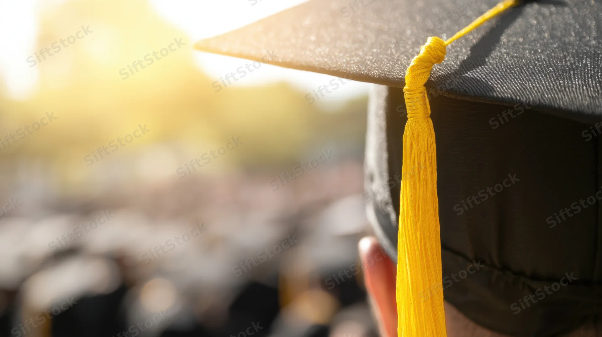 Close up of a black graduation cap with yellow tassel against a blurred outdoor ceremony background