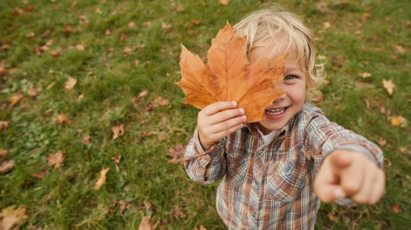 Smiling child holding autumn maple leaf over face while pointing forward in grassy yard