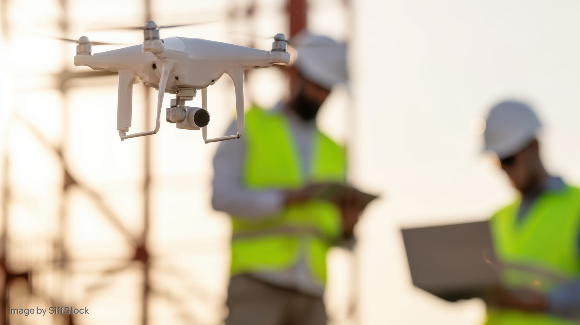 White drone hovering over construction site with workers in safety vests and helmets using tablets and laptops