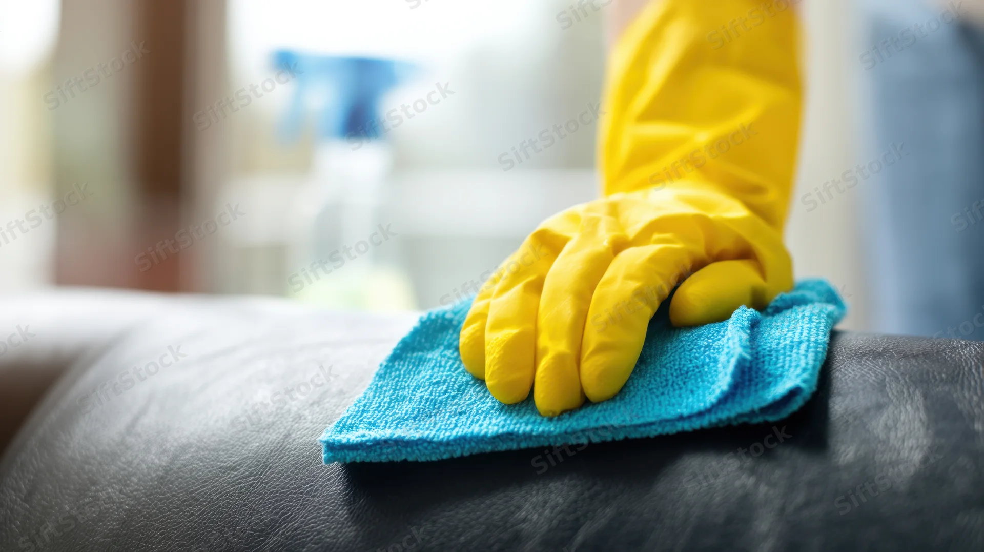 Hand in yellow rubber glove wiping leather sofa with blue cloth