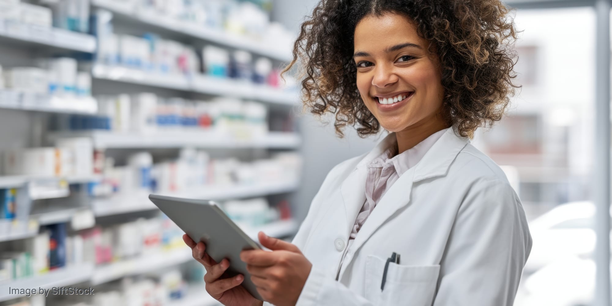 Smiling woman in white coat holding tablet in pharmacy aisle