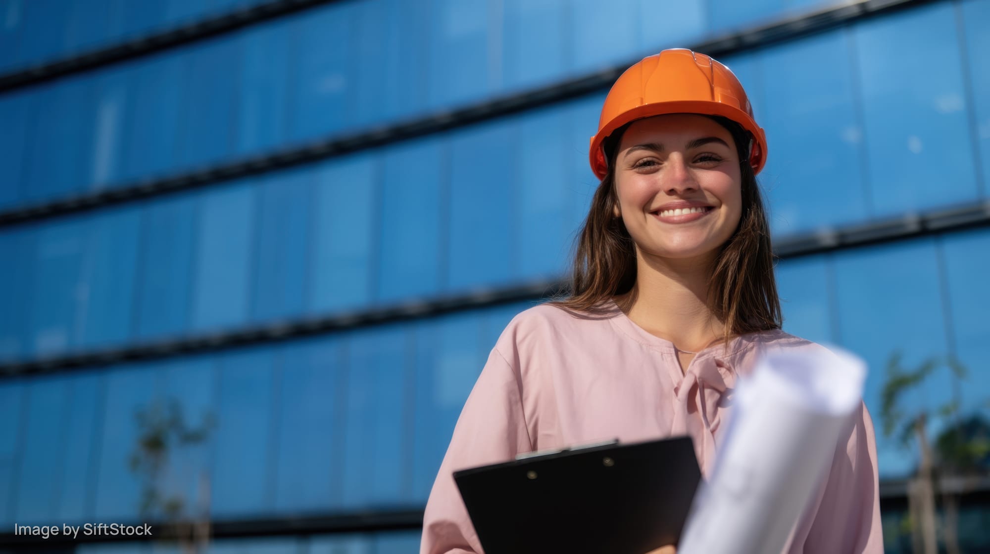 Smiling woman engineer in orange hardhat holding clipboard and blueprints outside modern glass building