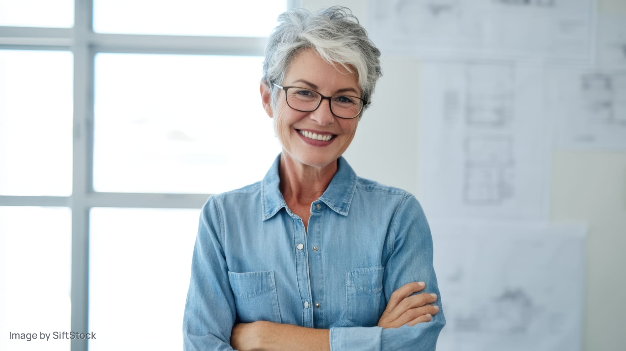 Smiling senior woman with arms crossed in an architect's office near a window with blueprints on the wall
