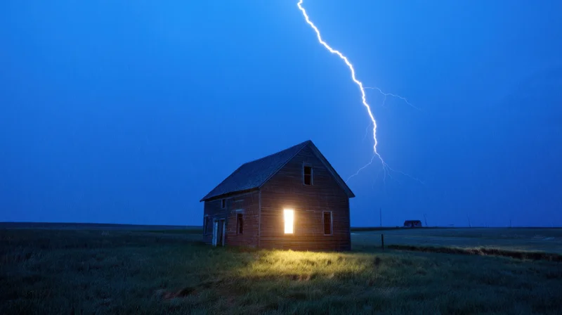 Weathered wooden house with illuminated window during lightning strike in rural field at night