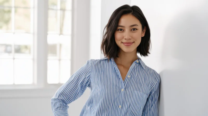 Asian woman smiling in striped blue shirt leaning against white wall near window