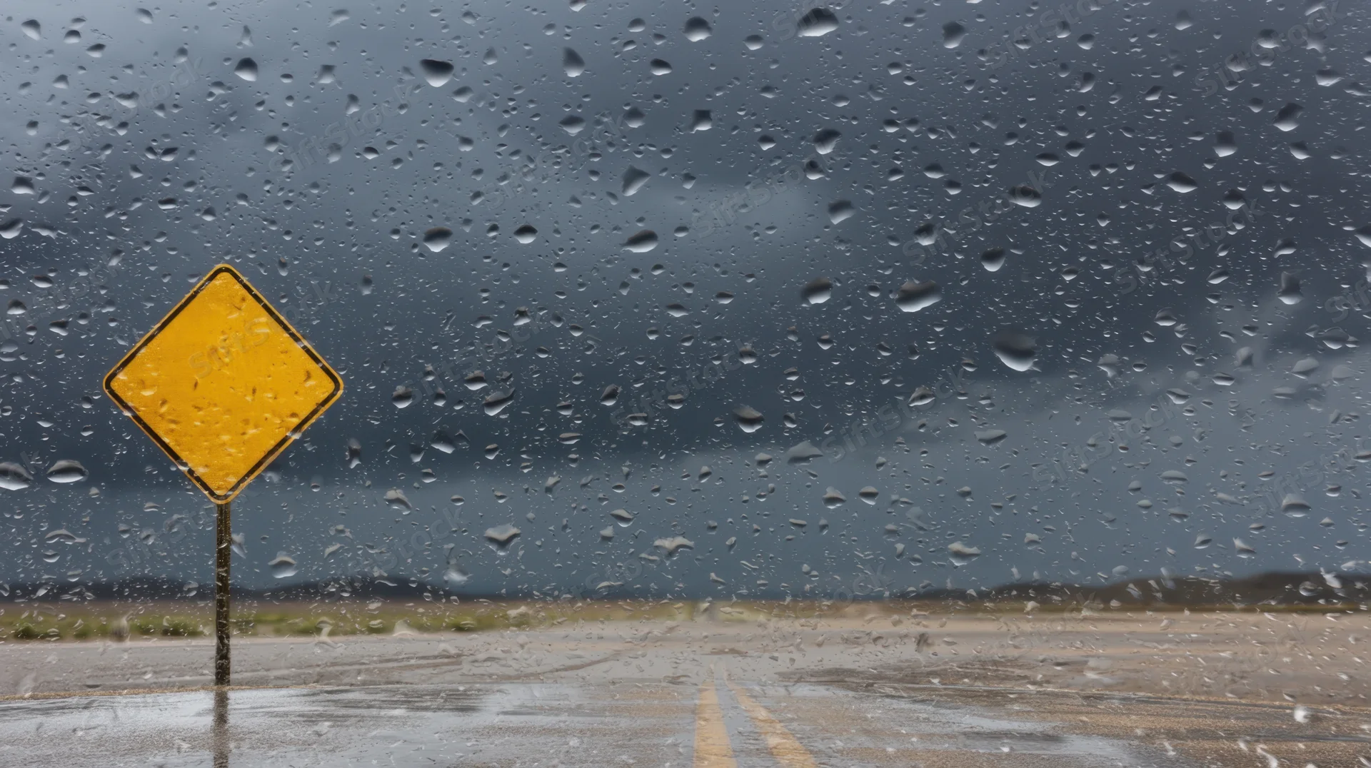 Yellow diamond road sign viewed through rain-covered windshield on wet highway
