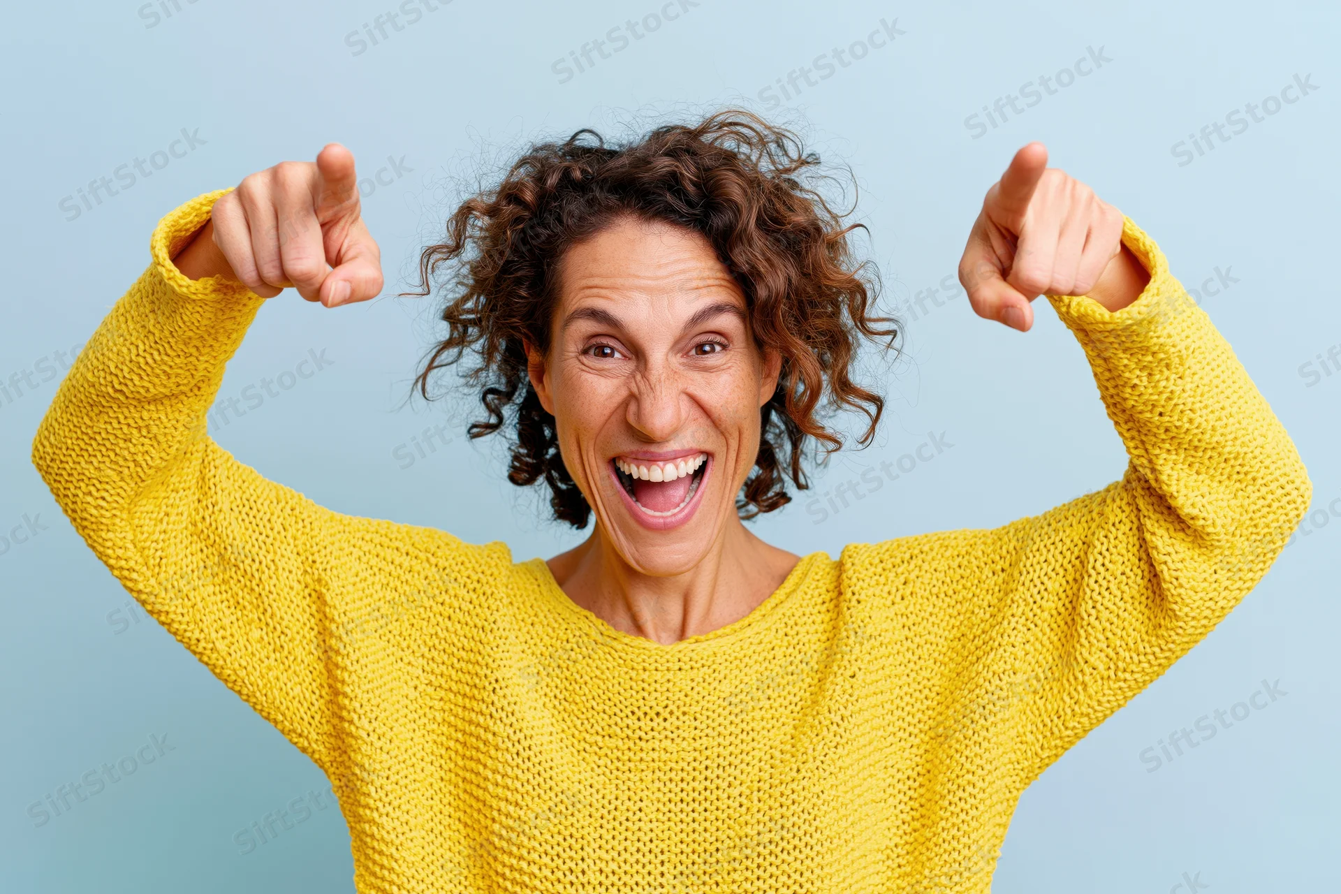 Woman in yellow knit sweater pointing with raised arms, smiling excitedly against blue background