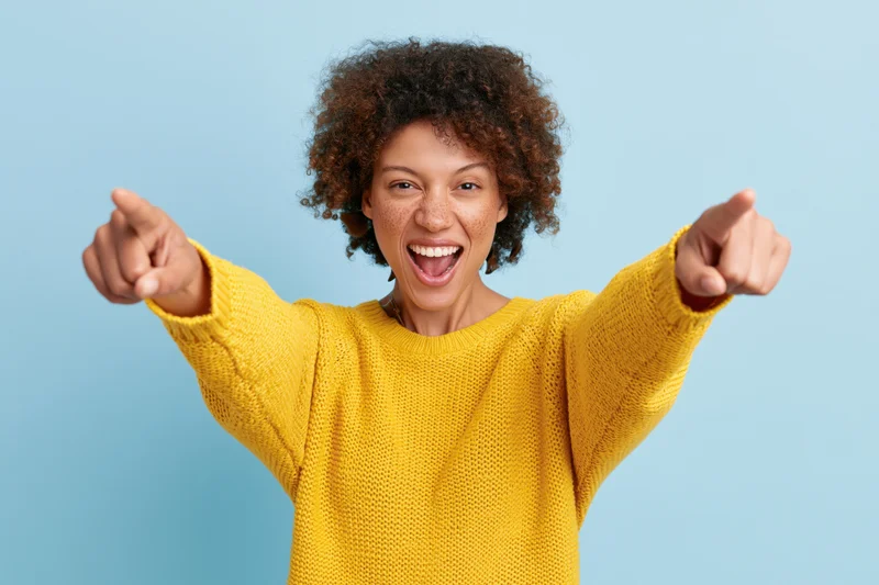 Woman in yellow knit sweater pointing with both hands while smiling widely against blue background
