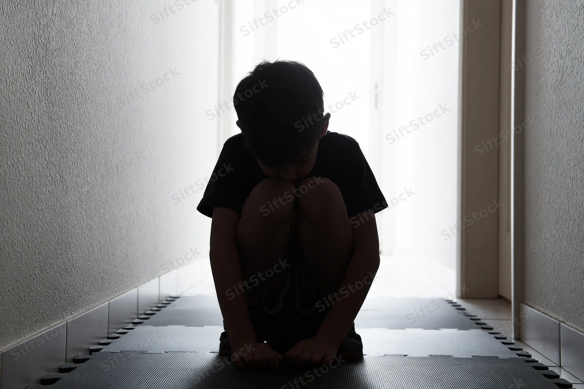 Child crouching on foam puzzle mat in dim hallway with light from doorway
