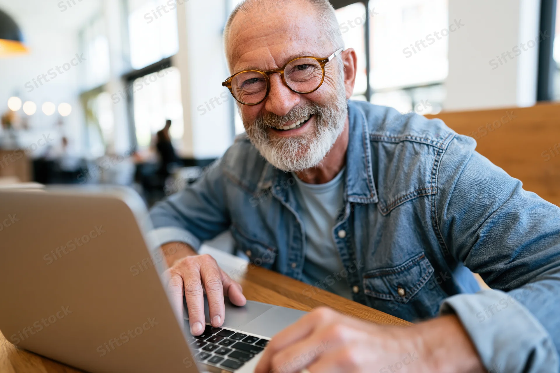 Senior man smiling while working on laptop at wooden desk in modern office space