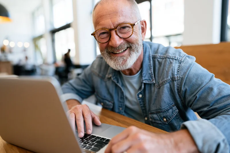 Senior man smiling while working on laptop at wooden desk in modern office space
