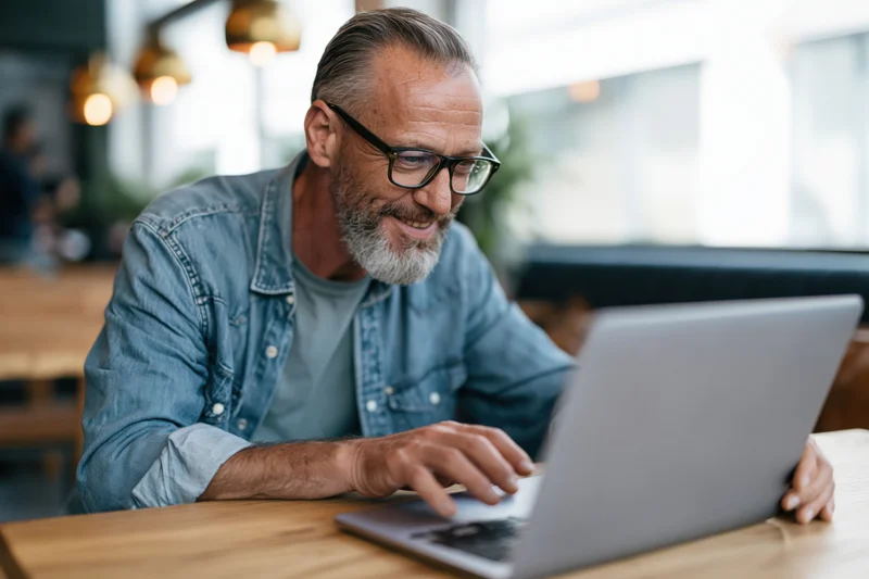 Smiling middle-aged man with gray beard and glasses working on laptop in cafe