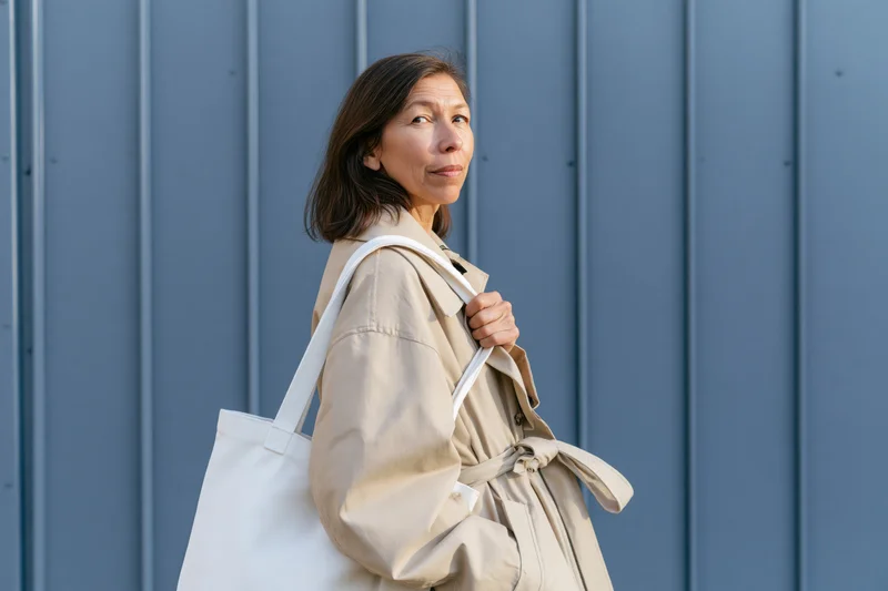 Woman in beige coat holding white tote bag standing against blue panel wall