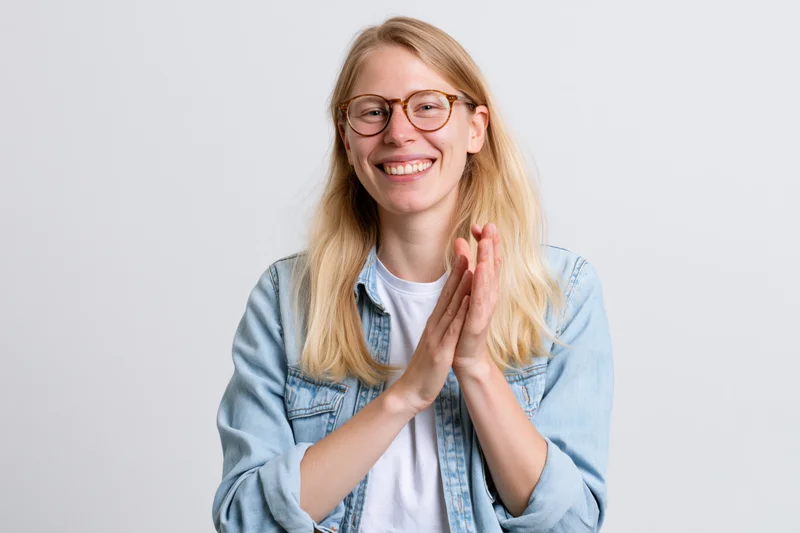 Young woman with long blonde hair and round glasses smiling and clapping hands in a studio setting