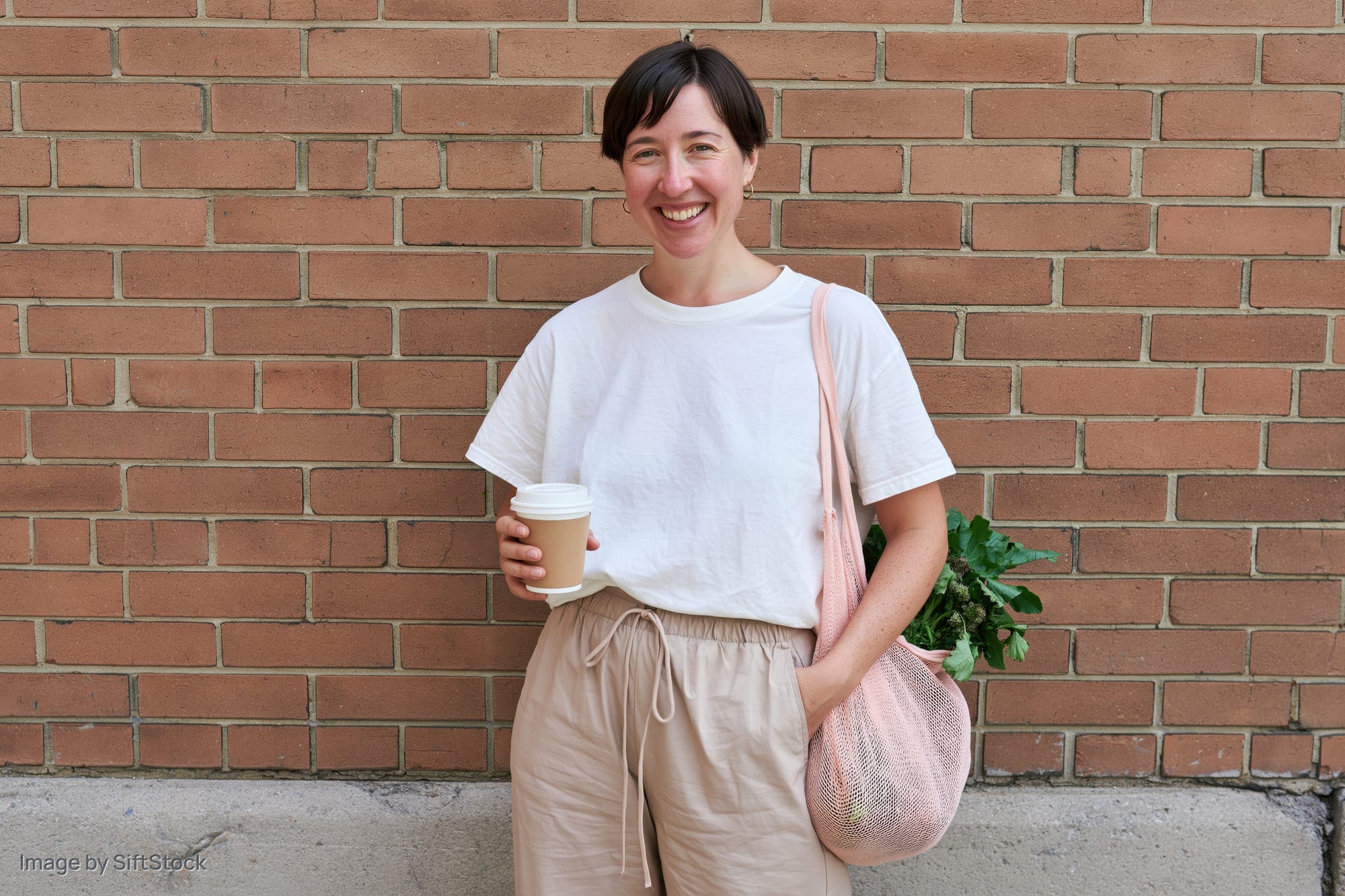 Woman smiling while holding coffee cup and carrying mesh bag with plant against brick wall