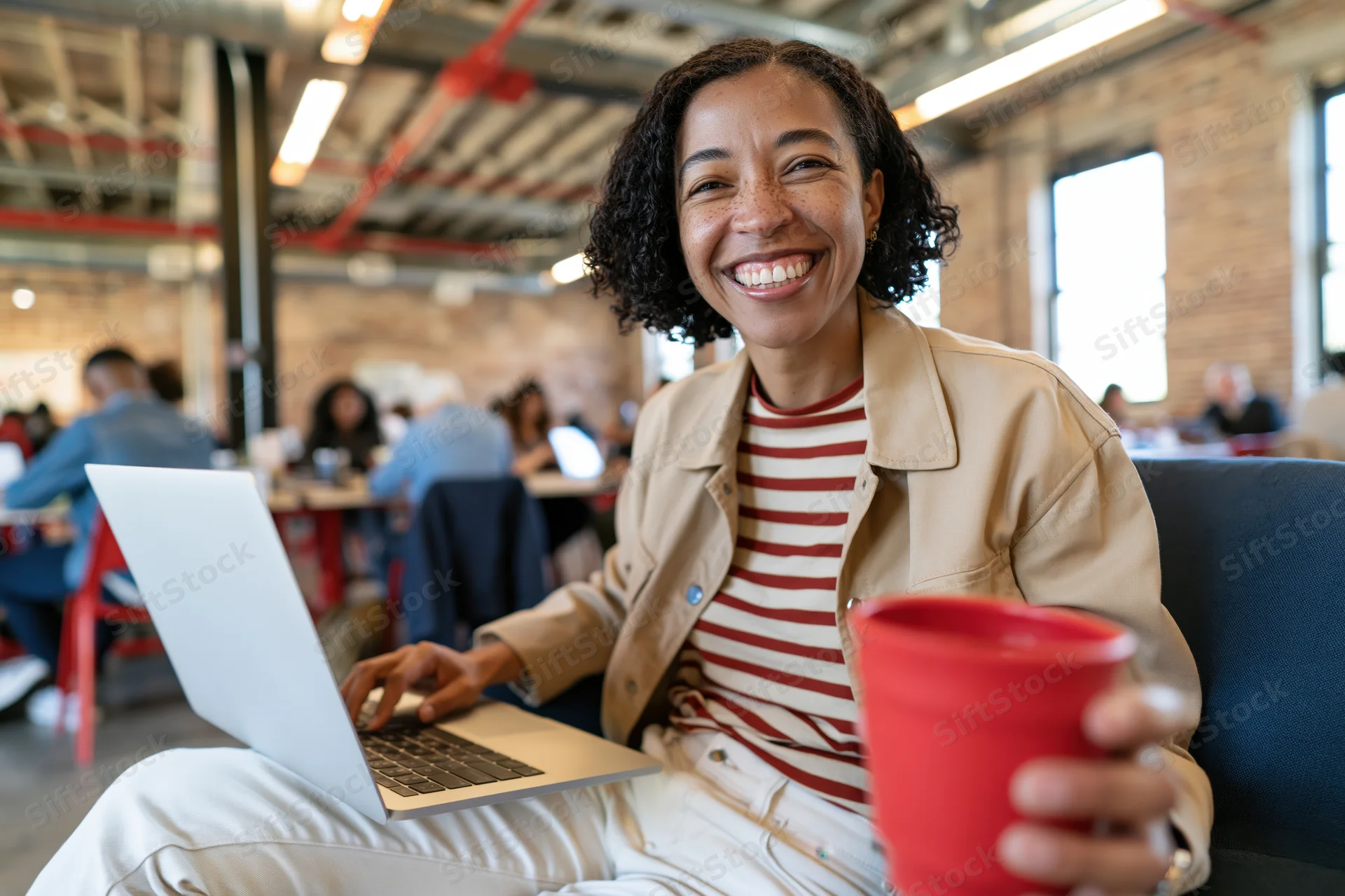 Woman smiling at camera while using laptop and holding coffee cup in modern cafe workspace