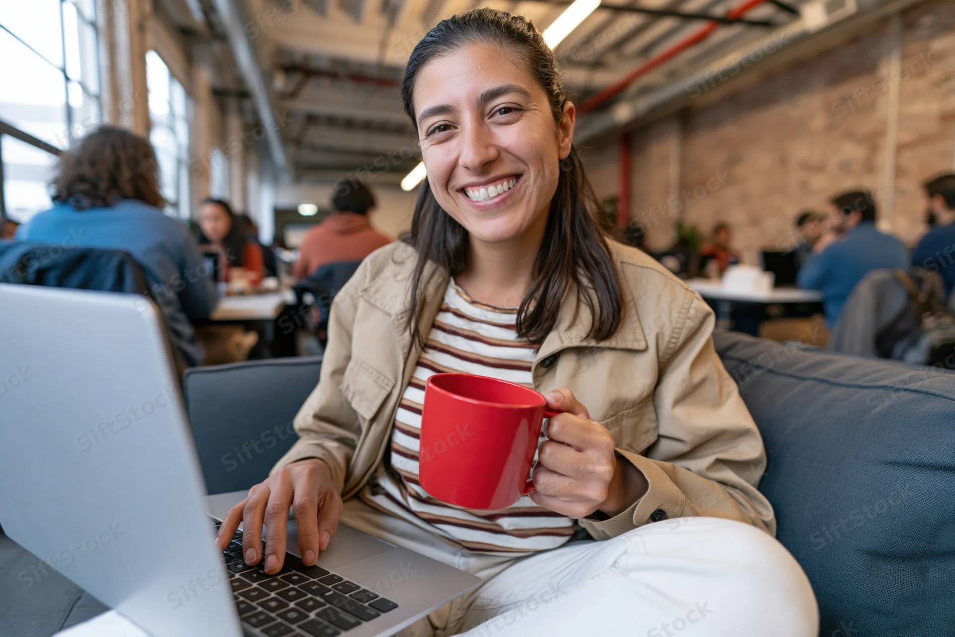 Woman smiling at camera while typing on laptop and holding red mug seated on couch in modern office space