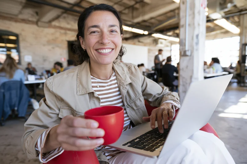 Woman smiling at camera while using laptop and holding red mug in open office space
