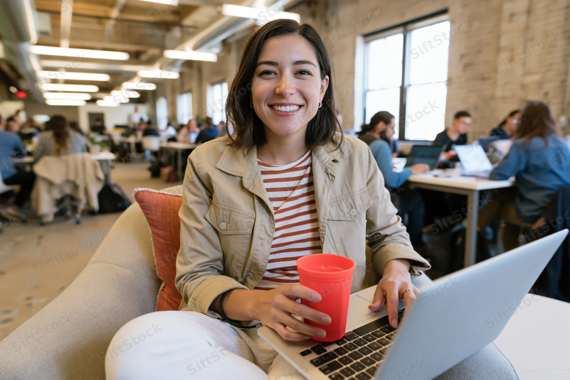 Woman smiling at camera seated on sofa with laptop in modern office workspace