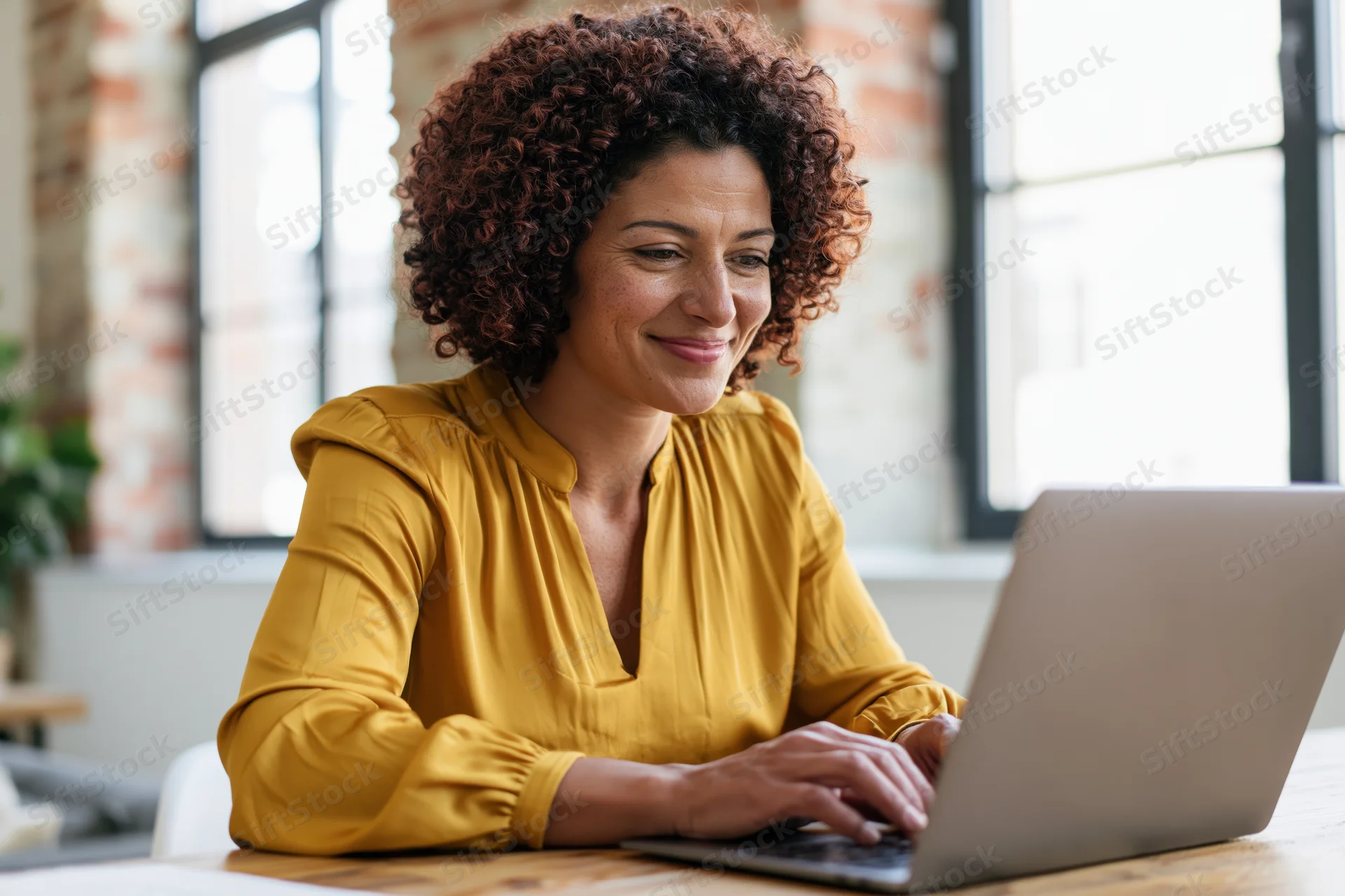 Woman working on laptop at wooden desk in modern office with brick walls and windows
