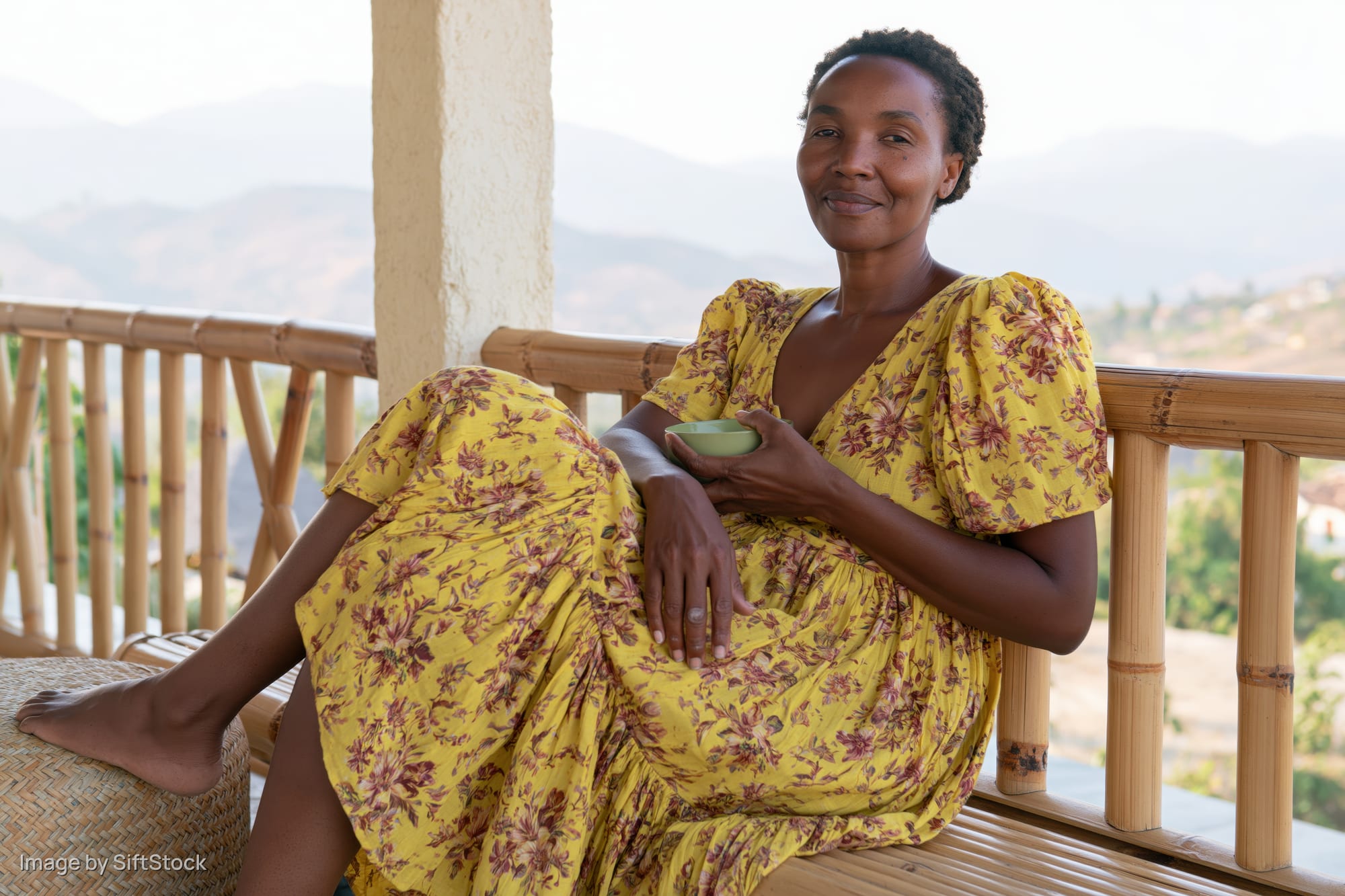 Woman in yellow floral dress sitting on balcony smiling with mountains in background