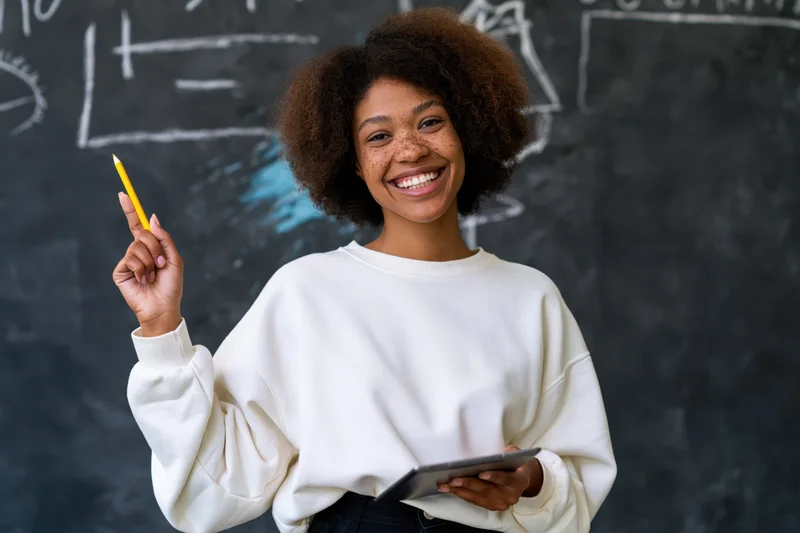 Young woman smiling while holding tablet and pencil in front of blackboard