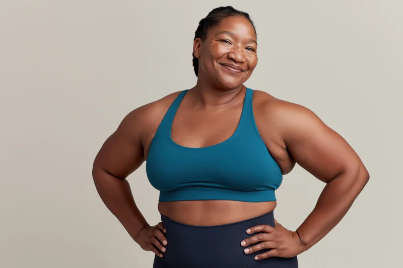 Plus size woman in teal sports bra and dark leggings posing with hands on hips in studio