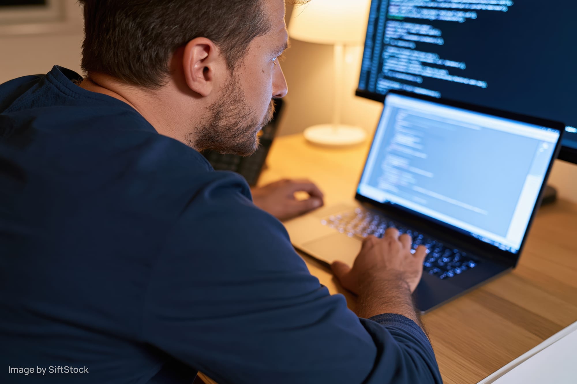 Man typing code on laptop at desk with monitor in home office setting