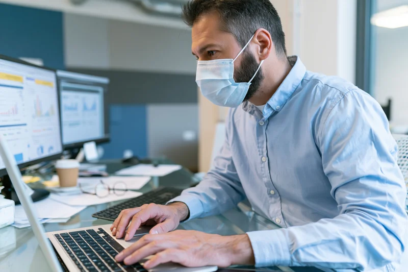 Man wearing face mask typing on laptop at office desk