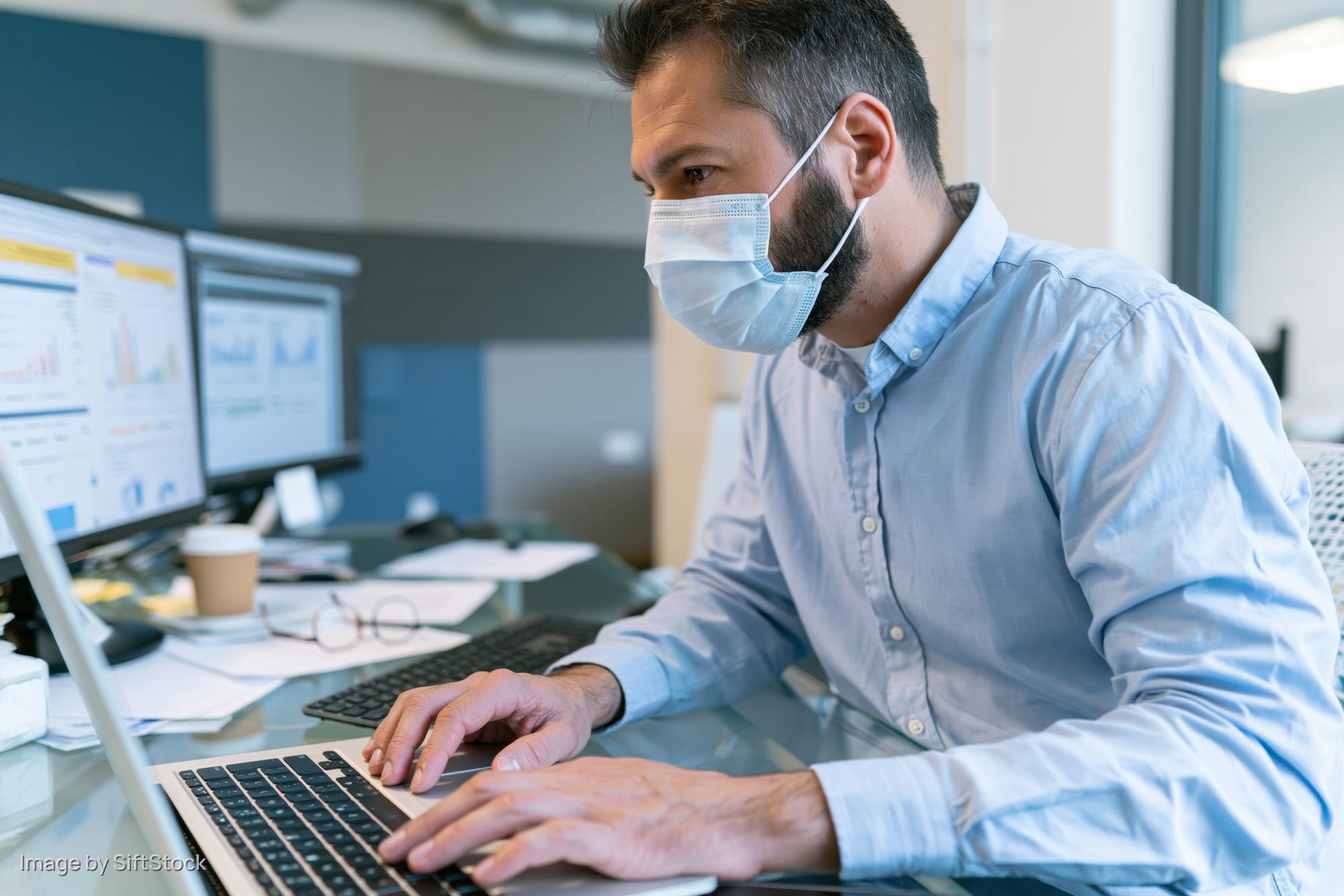Man wearing face mask typing on laptop at office desk