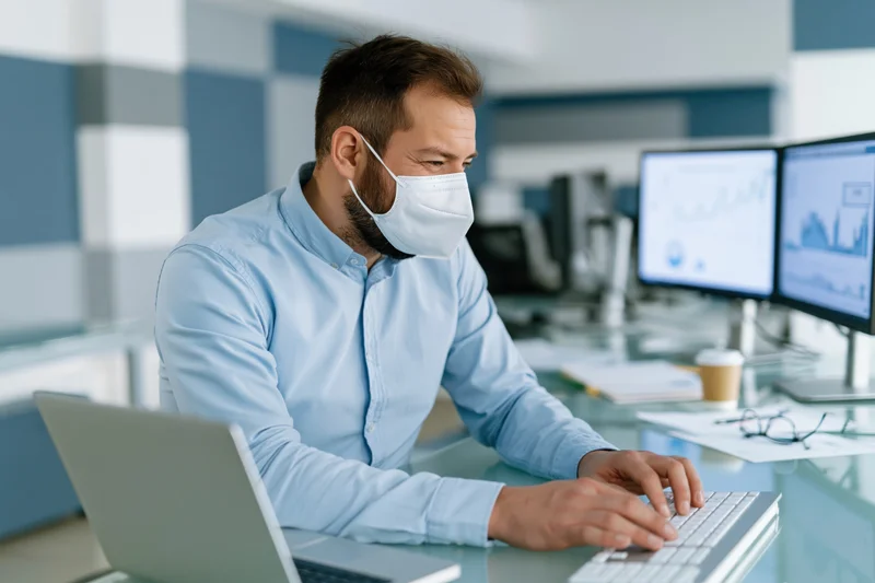 Man wearing face mask typing on keyboard at office desk with dual monitors