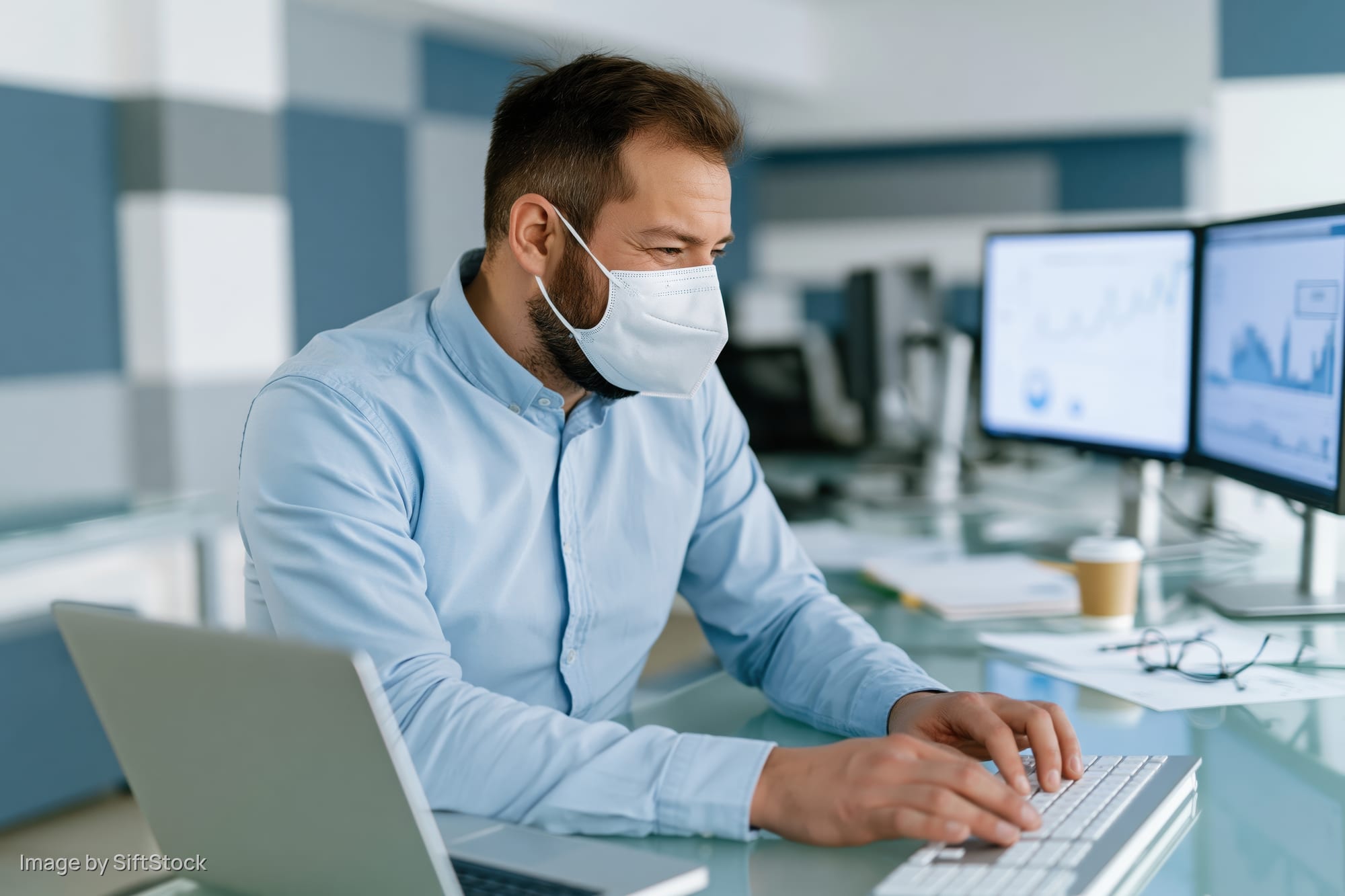 Man wearing face mask typing on keyboard at office desk with dual monitors