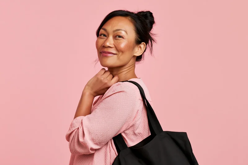 Young asian woman smiling softly in three-quarter pose with tote bag over shoulder against pink background